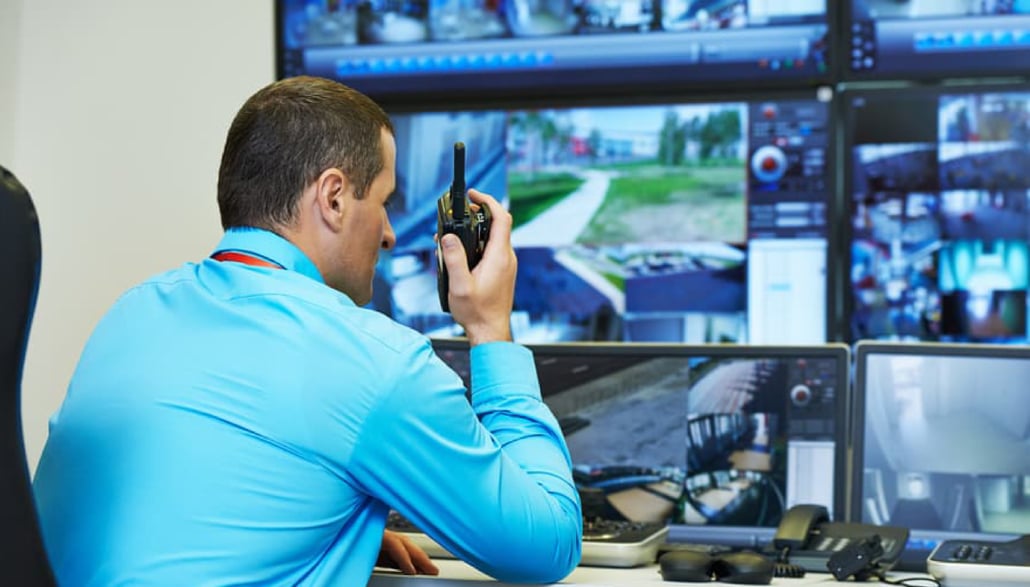 Man monitoring video surveillance on multiple screens and speaking into walkie talkie