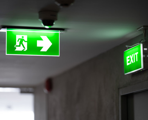 A green fire exit sign is placed on the ceiling along the dimly lit corridor and there is green exit sign on the exit door.