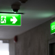 A green fire exit sign is placed on the ceiling along the dimly lit corridor and there is green exit sign on the exit door.